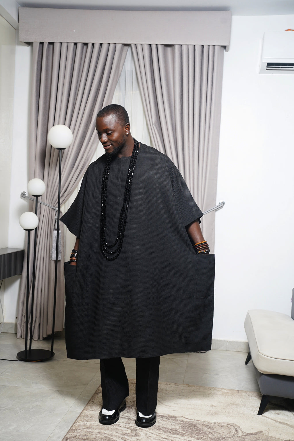 Model wearing black agbada with layered black beads holding carved butterfly staff in a contemporary indoor space