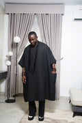Model wearing black agbada with layered black beads holding carved butterfly staff in a contemporary indoor space