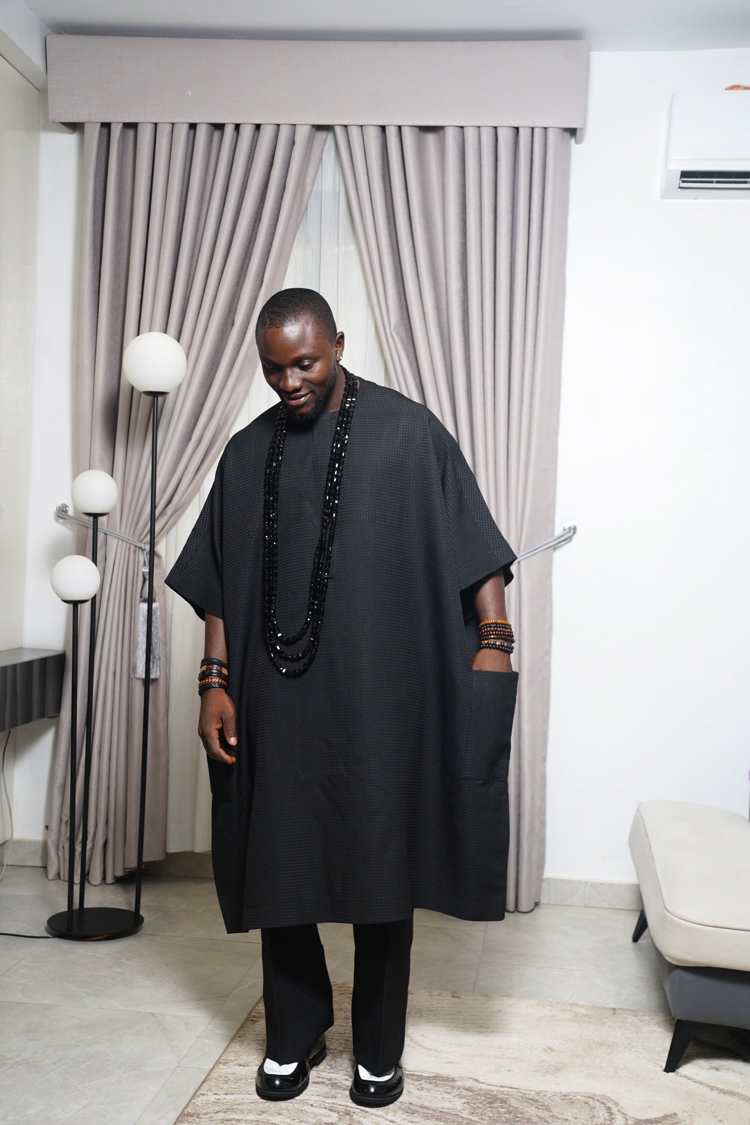 Model wearing black agbada with layered black beads holding carved butterfly staff in a contemporary indoor space