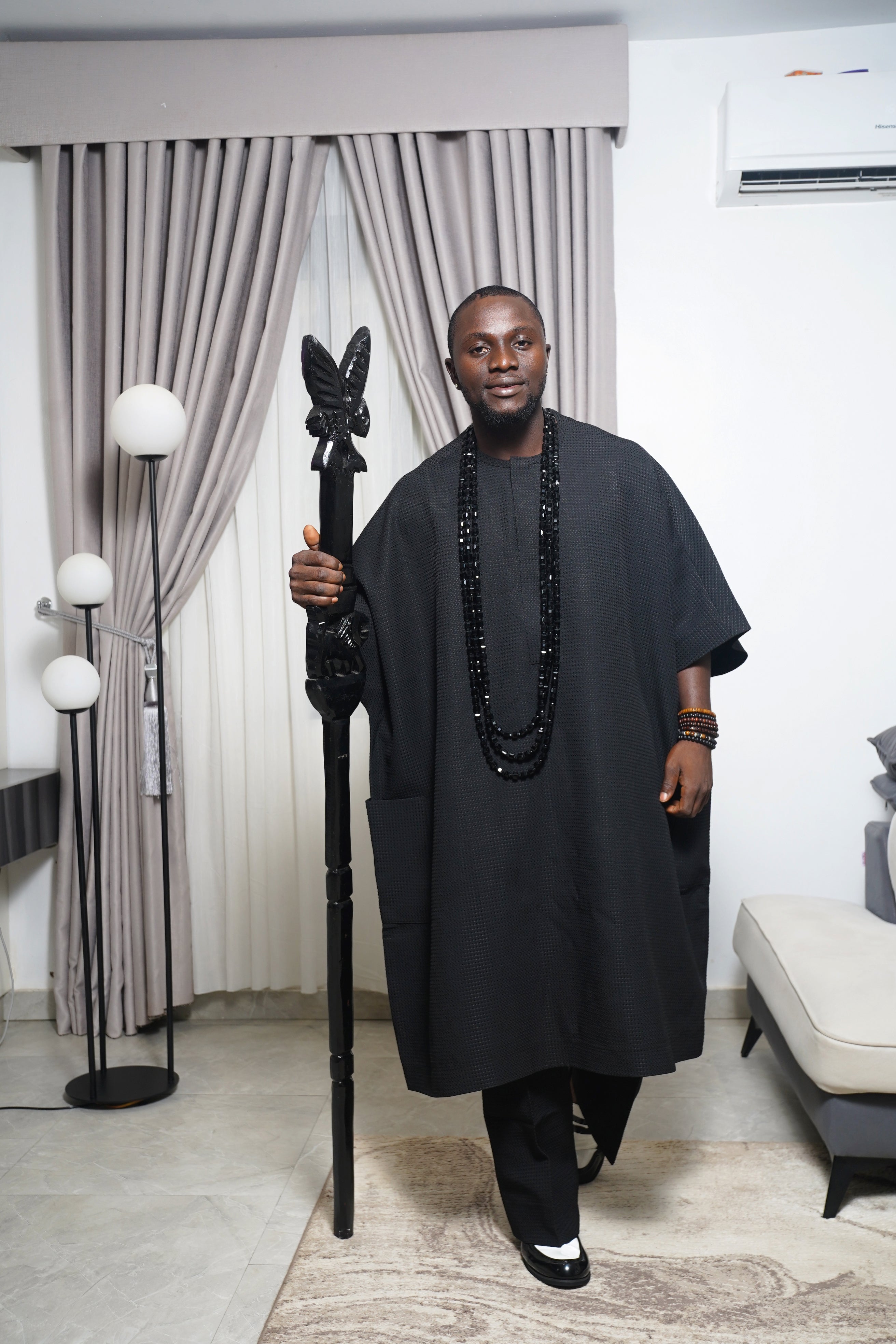Traditional black Igbo outfit styled with coral beads and red beaded staff, photographed indoors with modern décor