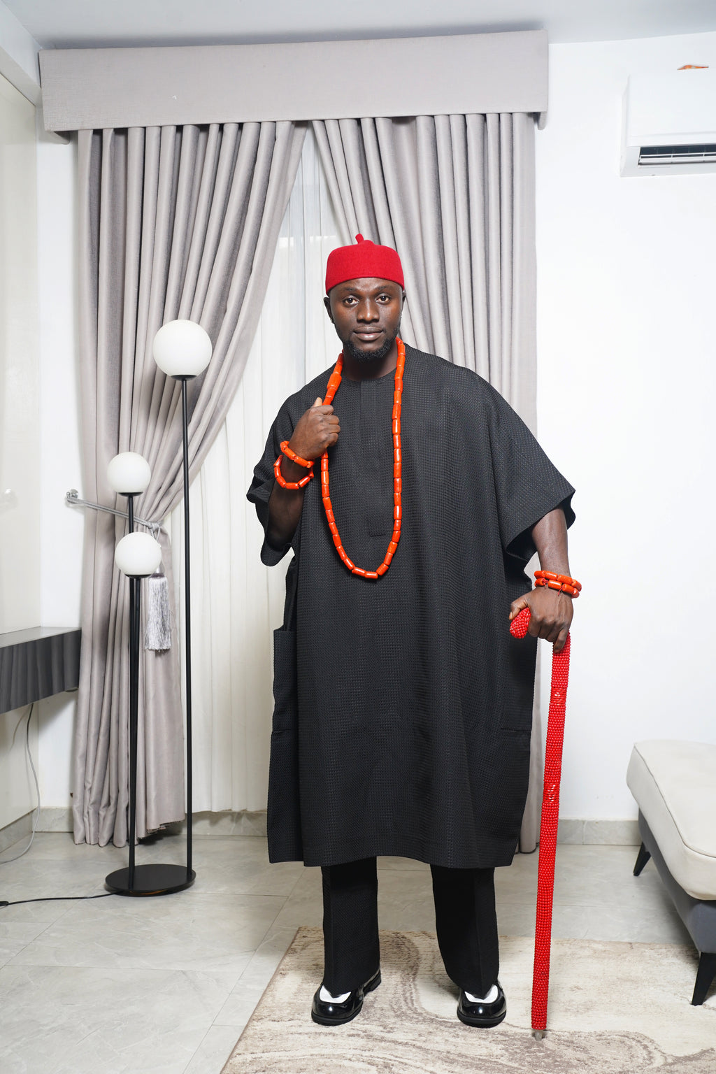 Traditional black Igbo outfit styled with coral beads and red beaded staff, photographed indoors with modern décor.