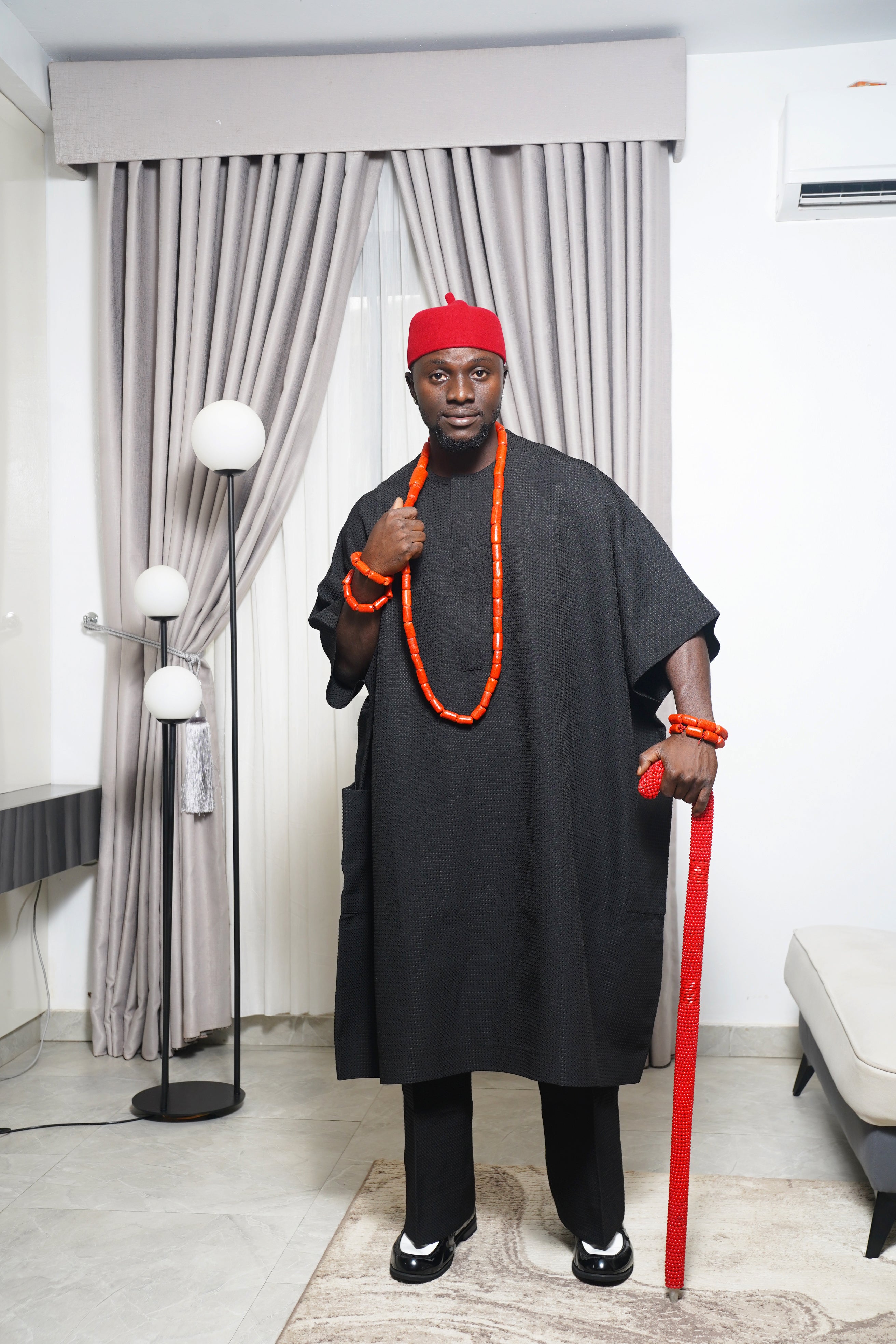 Traditional black Igbo outfit styled with coral beads and red beaded staff, photographed indoors with modern décor.