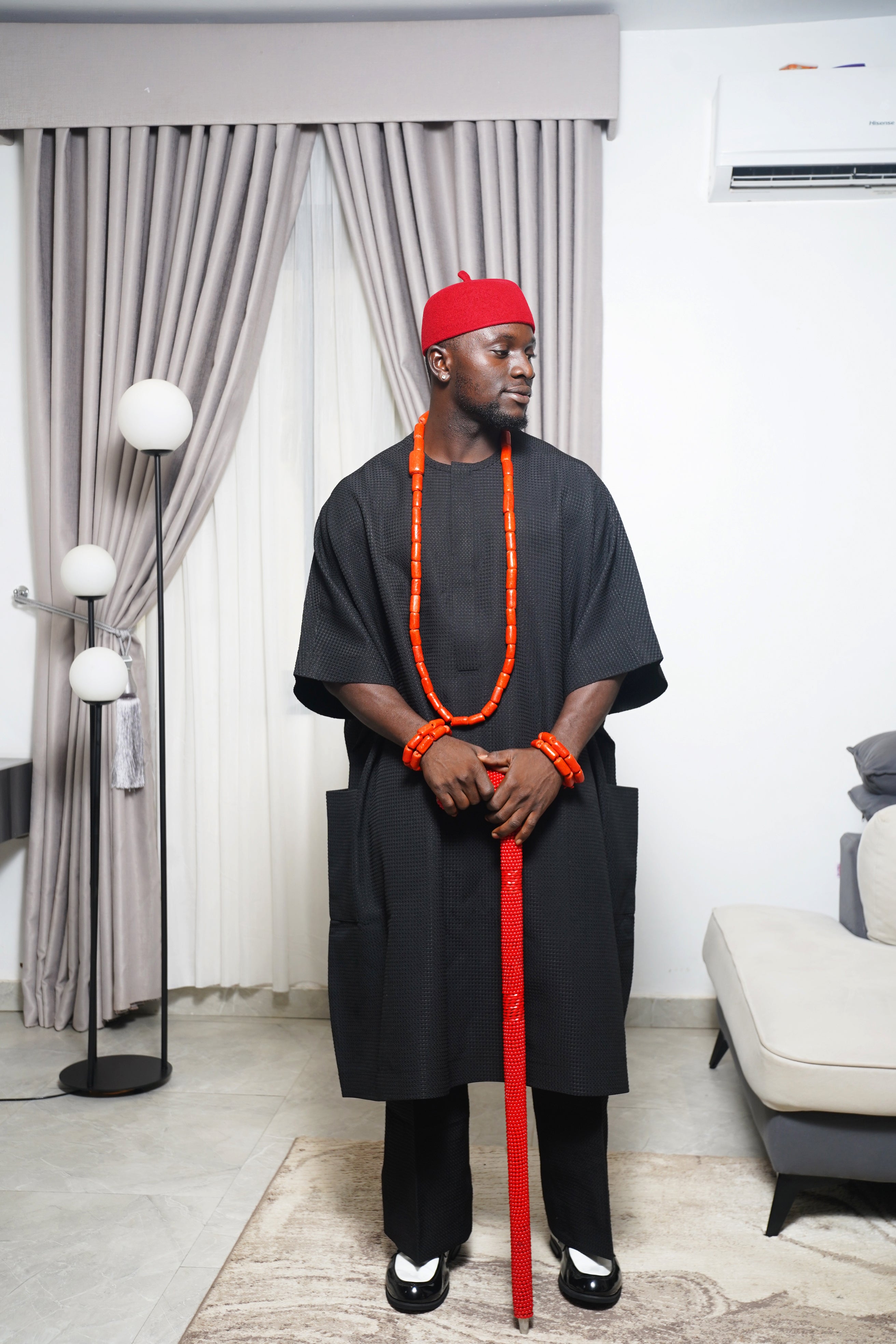 Model wearing black traditional Igbo attire with red cap, coral bead necklaces, and red beaded staff in a modern indoor setting