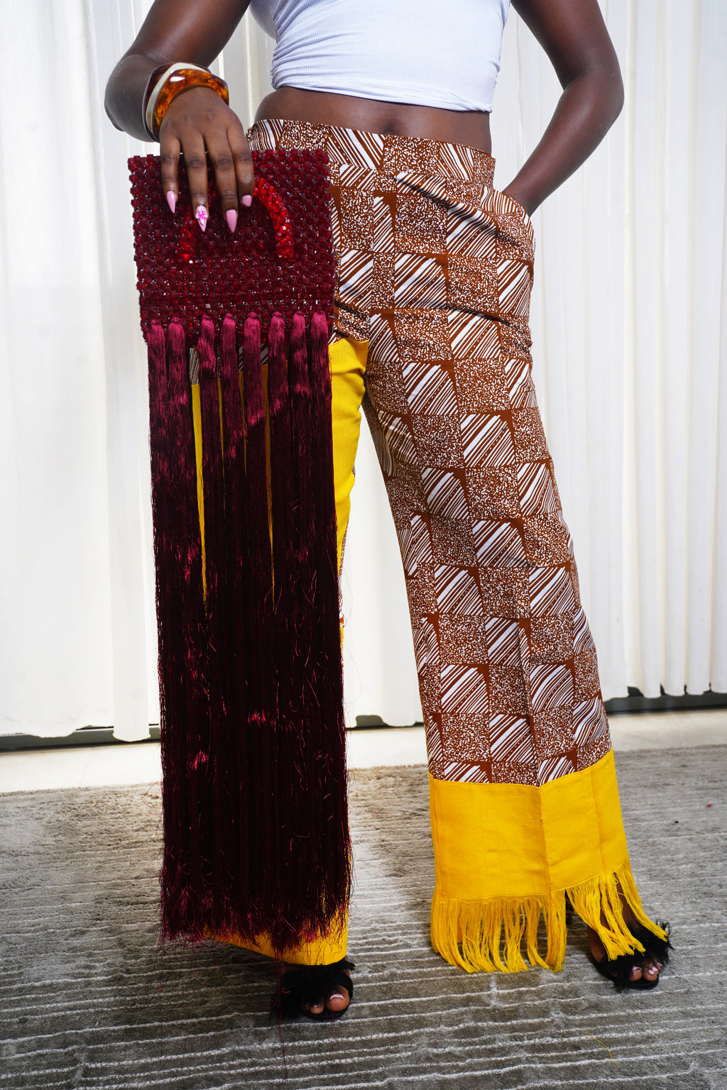 Model holding red beaded fringe handbag while wearing brown African print pants with yellow fringed cuffs.