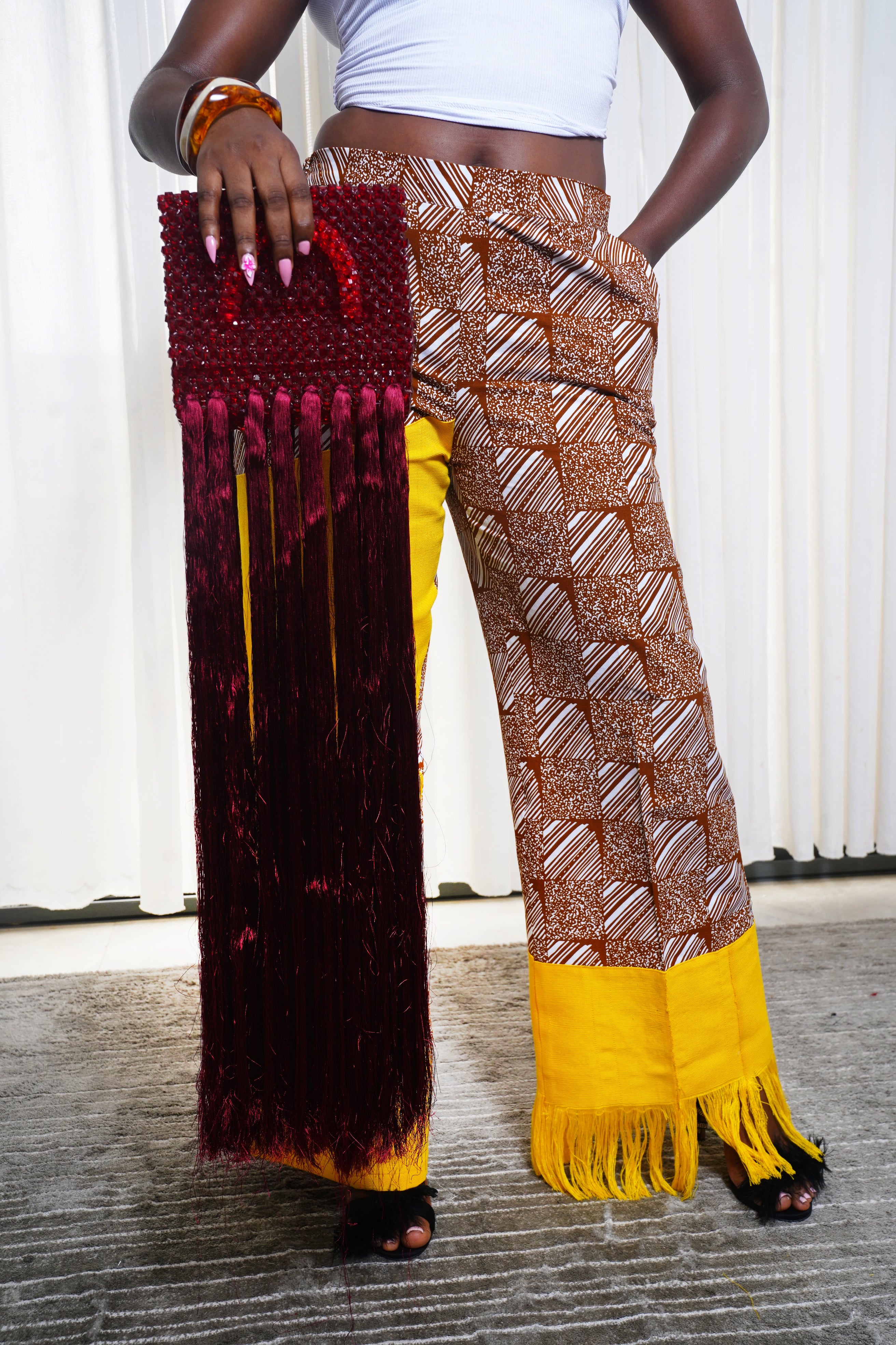 Model holding red beaded fringe handbag while wearing brown African print pants with yellow fringed cuffs.